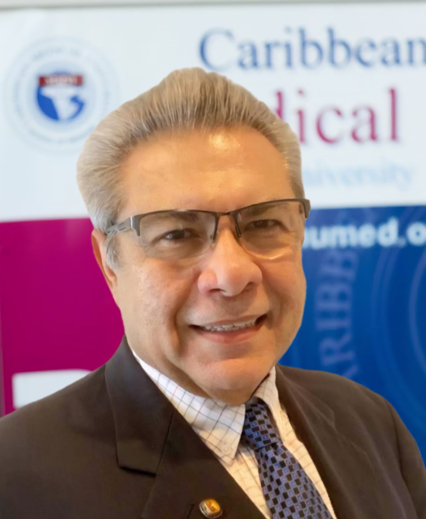Man in a suit with a blue tie smiling, standing in front of a Caribbean Medical University banner.