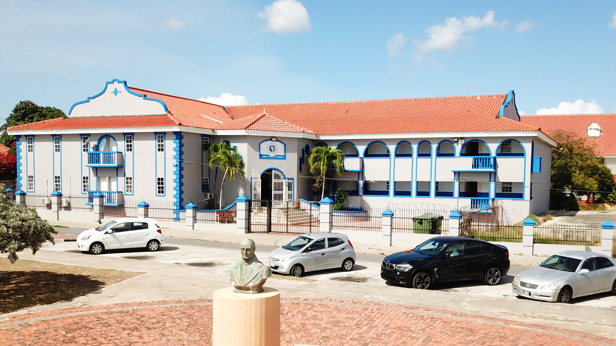 Yellow building with red-tiled roof, arched windows, and green lawn, suggesting a university dormitory in Curaçao.